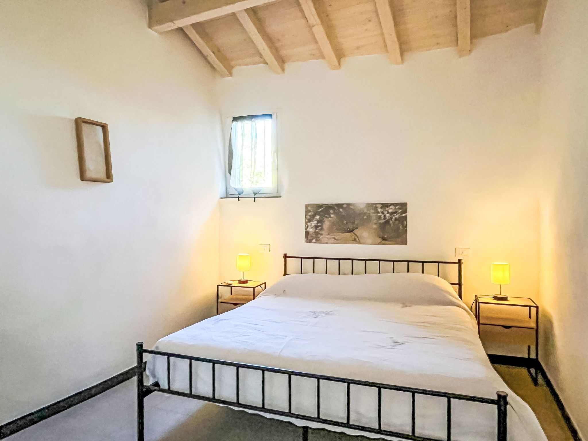 Une chambre à coucher avec un lit en métal, deux lampes de chevet, une fenêtre avec des rideaux et un tableau encadré au mur.