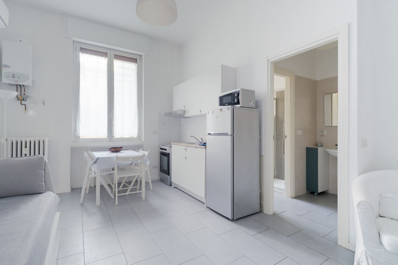 A compact kitchen with a refrigerator, stove, and microwave. There is a dining table with chairs and a window with blinds.