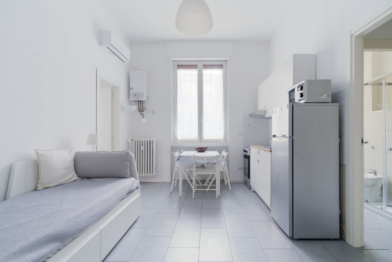 A compact, modern room with a bed, a dining table, a refrigerator, and a window.
