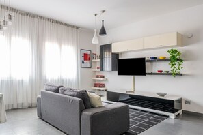 Een moderne woonkamer met een bank, een flatscreentelevisie, een salontafel en een keukenhoek met kasten en een plant.