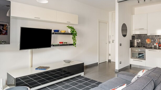 A modern living room with a flat-screen TV, a white entertainment unit, a grey sofa, and a kitchen area with white cabinets and a stove. - GuestHost - Charming apartment of about 50 square meters, perfect for two people, located on the first floor of a building with a lift (NOT suitable for disabled people).The property is located in the heart of the historic center of Verona, a few steps f (Verona)