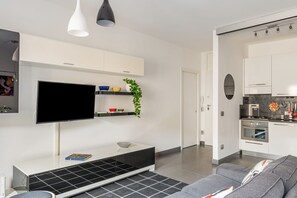 Ett modernt vardagsrum med en platt-TV, en vit möbel för underhållningssystemet, en grå soffa och en köksdel med vita skåp och en spis.