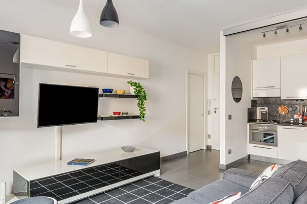A modern living room with a flat-screen TV, a white entertainment unit, a grey sofa, and a kitchen area with white cabinets and a stove.