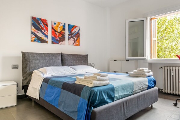 A modern hotel room with a large bed, a window with a view of trees, and abstract art on the wall.