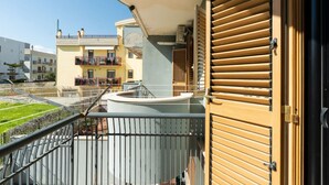 Un balcon doté d’une balustrade en métal, une porte avec des volets à persiennes horizontales et une vue sur les bâtiments voisins.