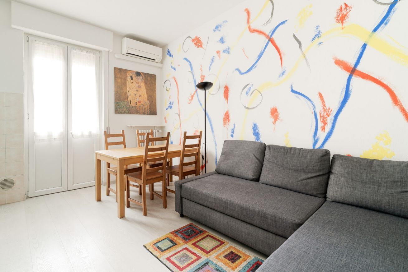 Een woonkamer met een grijze bank, een houten eettafel, stoelen en een kleurrijk vloerkleed.