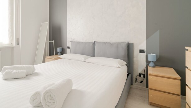 A neatly made bed with white linens, a bedside table with a lamp, and a nightstand with a lamp. - GuestHost - Gelsomini Apartment Milano (Milano)