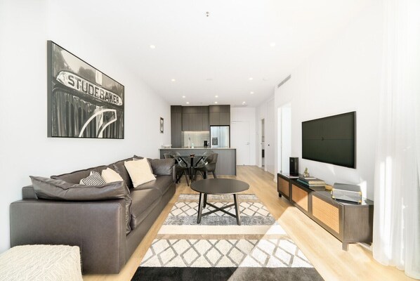 Una sala moderna con un sofá, una mesa de centro y un televisor. Se ve una cocina al fondo con armarios y una zona de comedor.