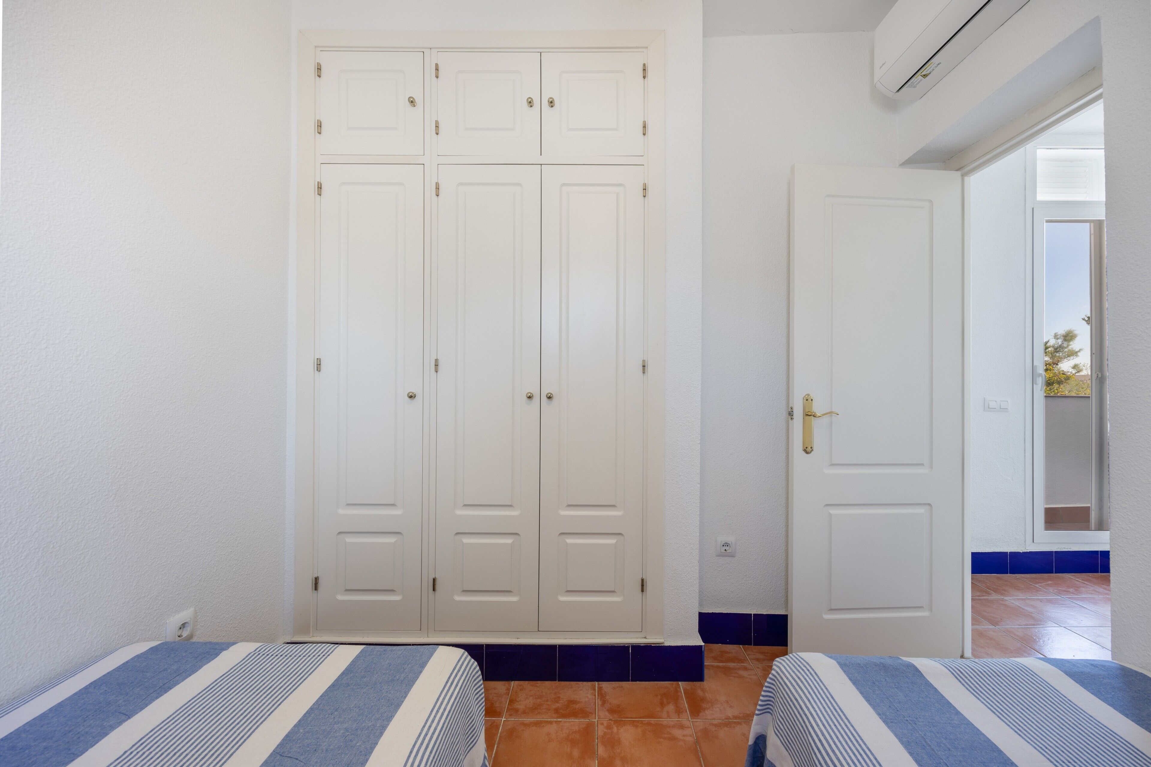 A room with two beds, a wardrobe, a door, and a view of the outside.