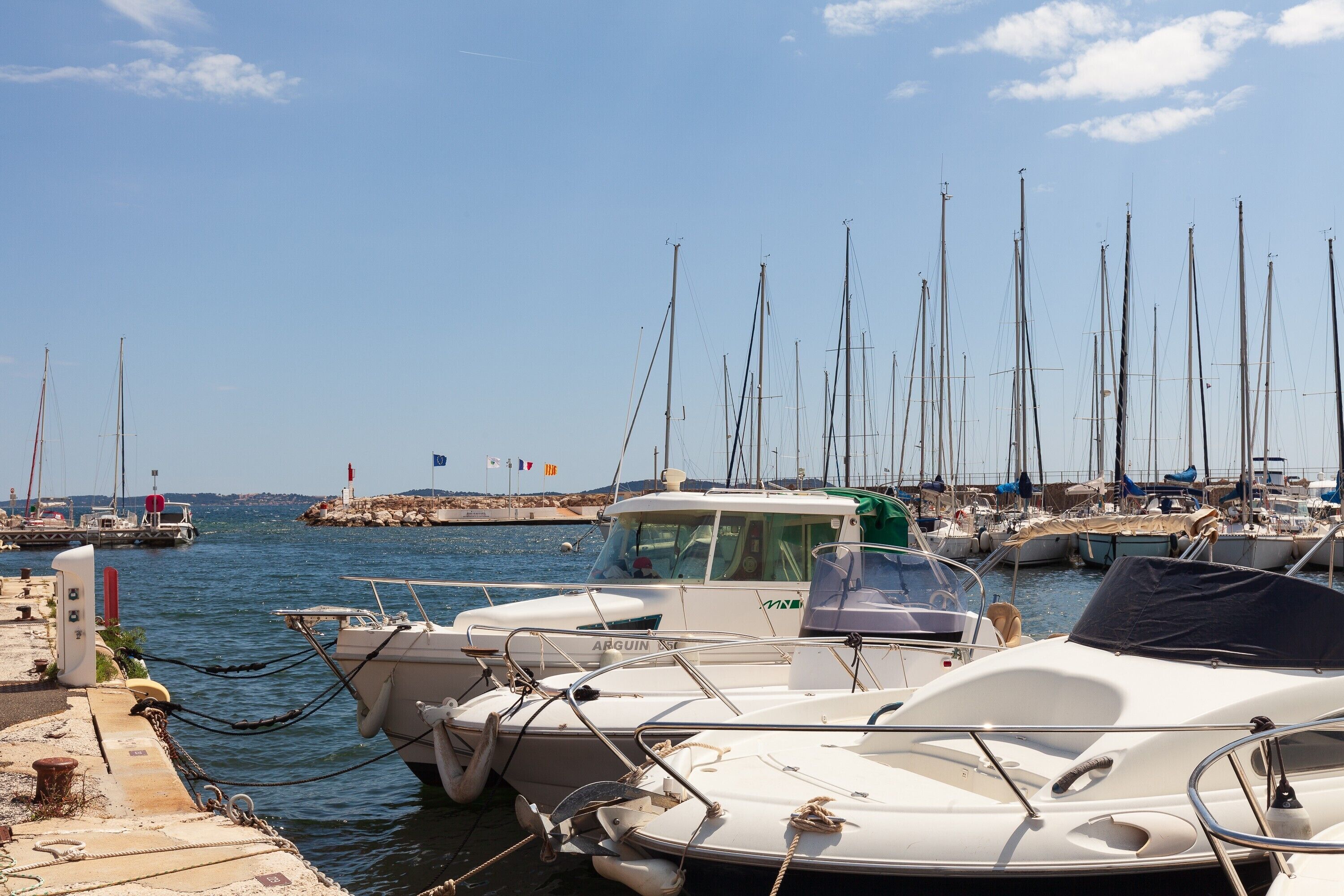 Un port de plaisance où sont amarrés de nombreux voiliers, une côte rocheuse en arrière-plan et un ciel dégagé.