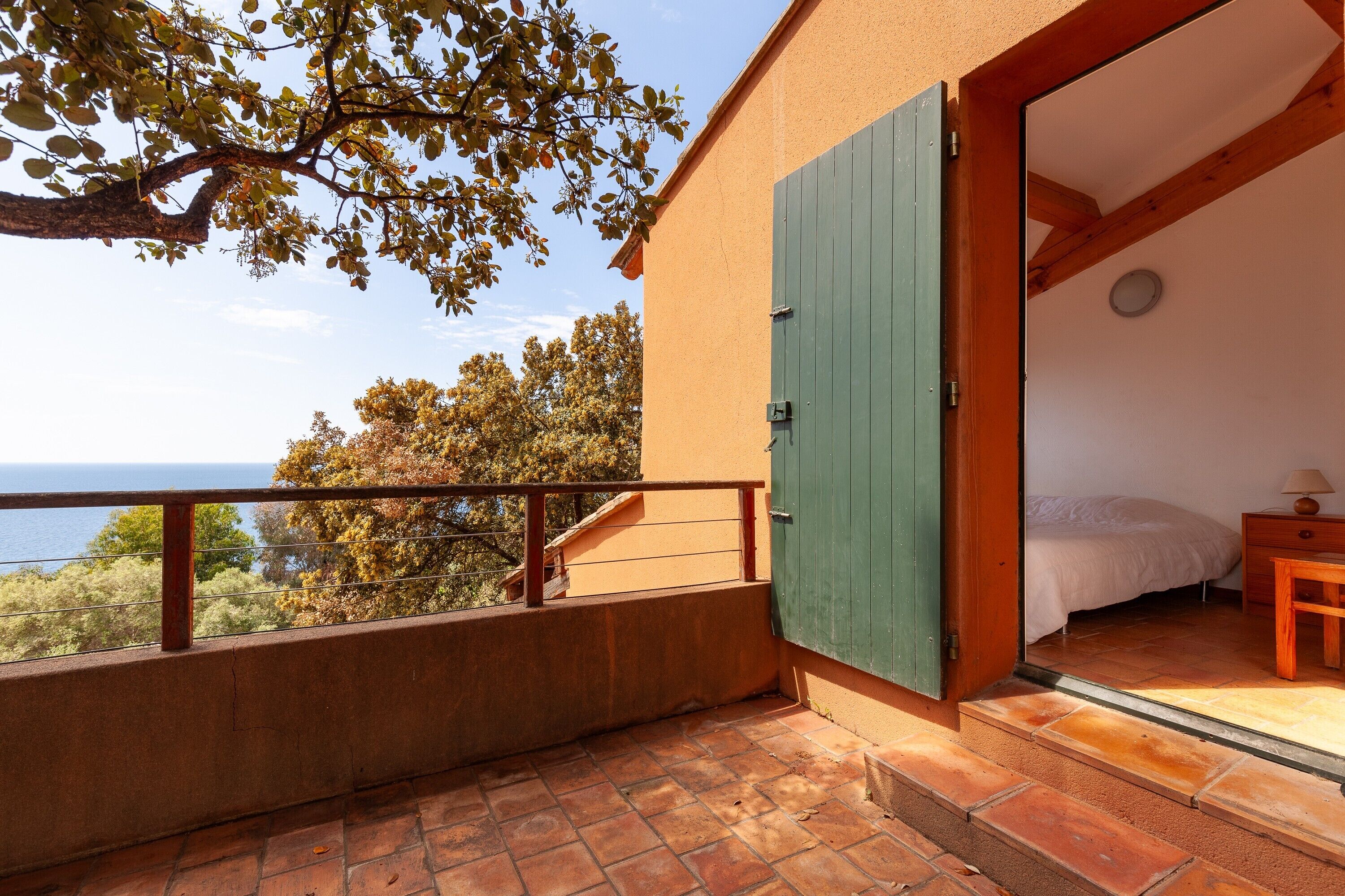 A balcony with a view of the sea, a bedroom with a bed and a nightstand, and a wooden door.