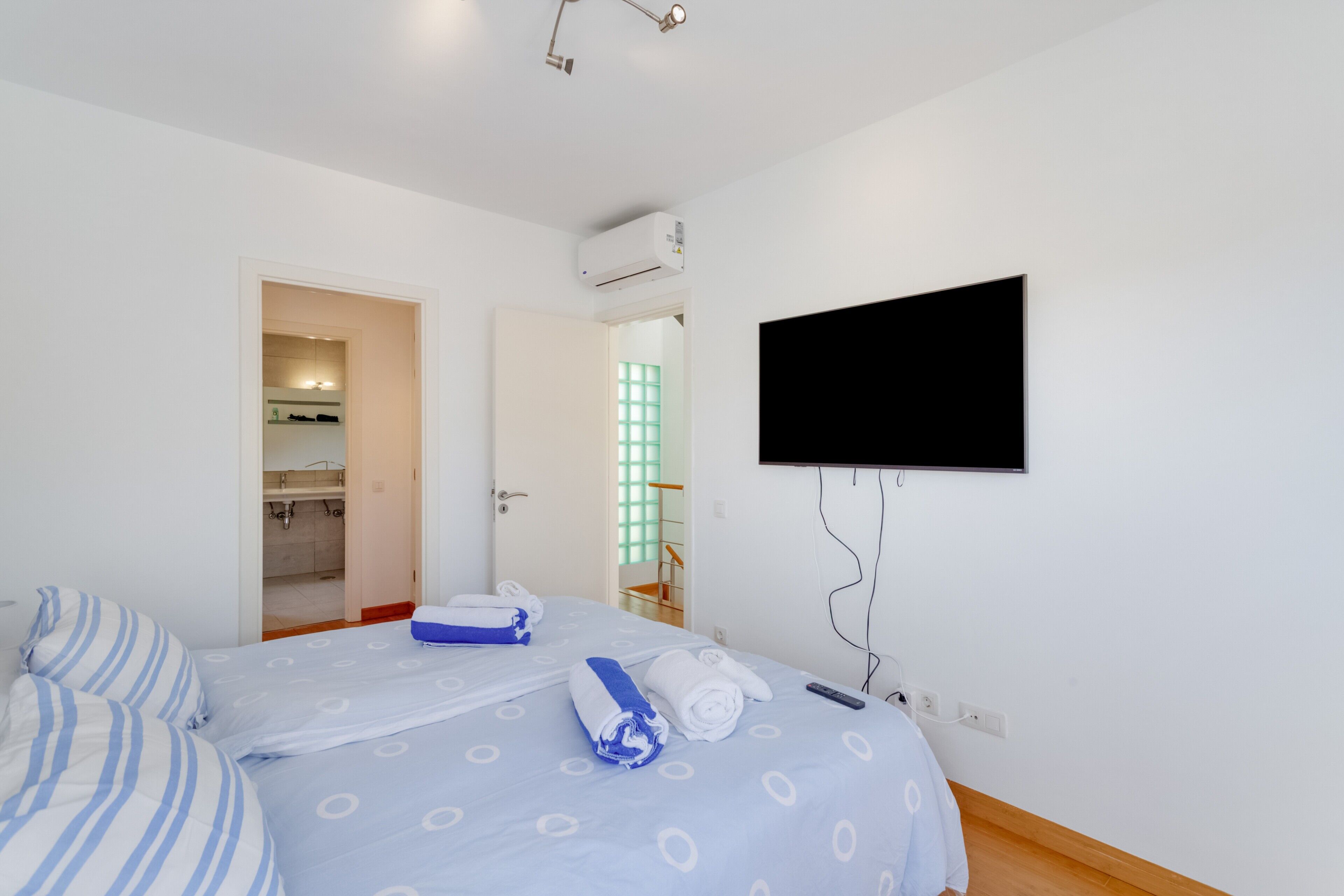 Habitación de hotel con cama, televisor montado en la pared, equipo de aire acondicionado y baño visible a través de una puerta abierta.