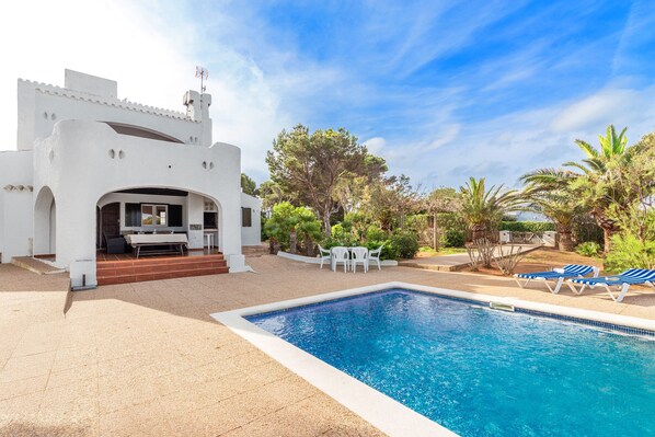 Une piscine avec des chaises longues, un espace patio et un bâtiment blanc doté d’un espace salon abrité.