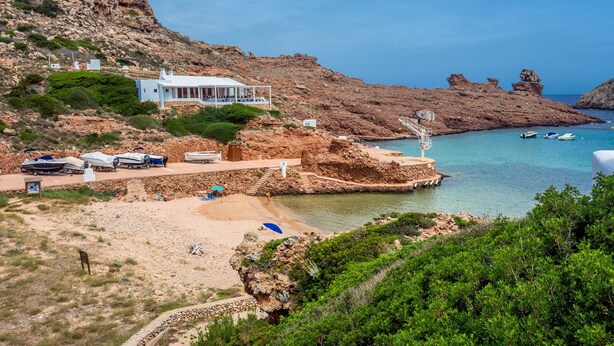 A coastal property with a white building, a beach, rocky cliffs, and boats in the water.
