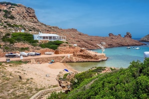 Une propriété située sur la côte, avec un bâtiment blanc, une plage, des falaises rocheuses et des bateaux sur l’eau.