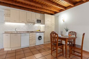 Uma cozinha com armários de madeira, micro-ondas, máquina de lavar louça e máquina de lavar roupa. Há uma mesa de jantar com cadeiras e um pequeno vaso com planta.