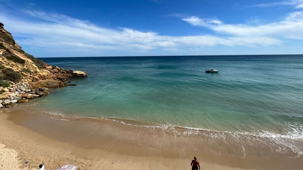 A beach with clear water, a rocky cliff, and a person walking on the sand.