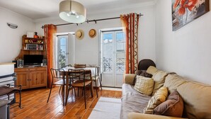 A living room with a sofa, TV, wooden cabinet, dining table, chairs, and a window with curtains.