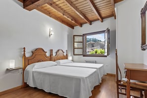 A bedroom with two beds, a wooden ceiling, a window with a view, and a desk.