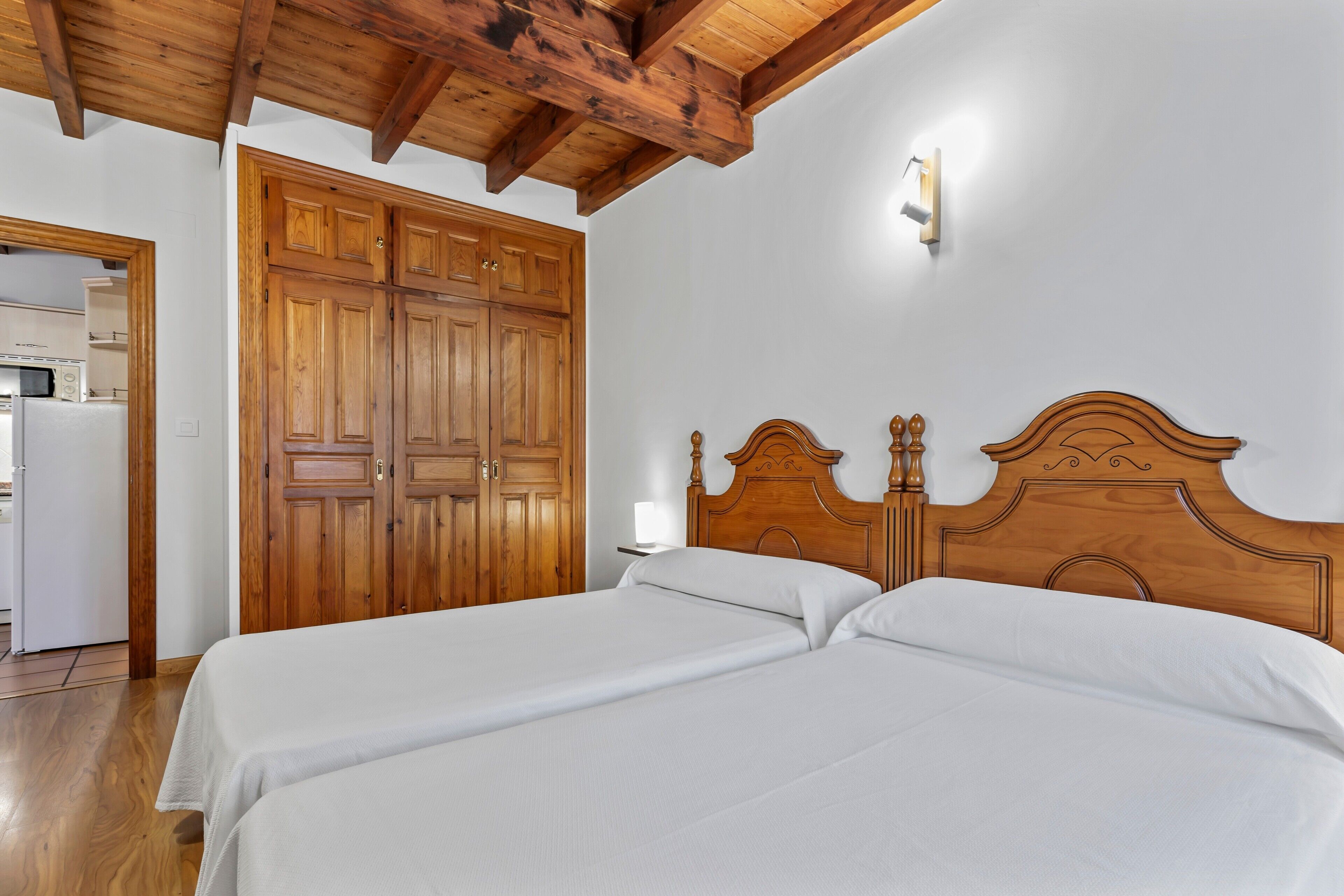 A bedroom with two beds, wooden wardrobe, and a kitchen visible through an open door.