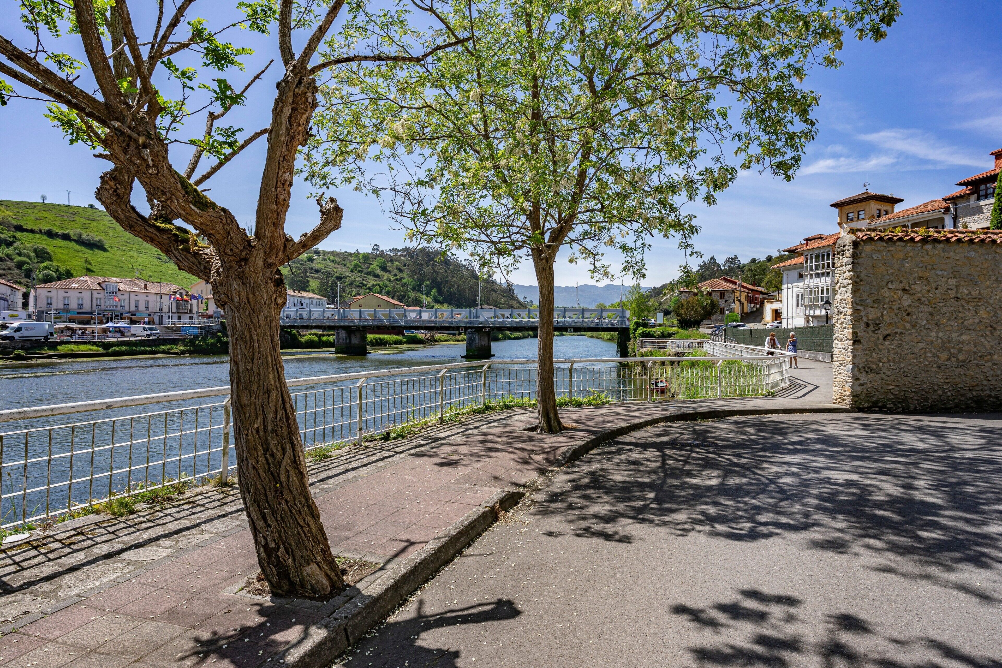 A riverside walkway with a stone wall, a bridge, and buildings in the background.
