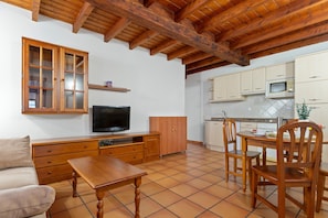 A compact living space with a wooden ceiling, a television, a dining table with chairs, a sofa, and a kitchen area with cabinets and a microwave.