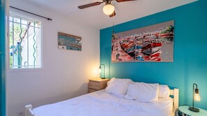 A bedroom with a bed, a ceiling fan, a window with bars, a wall painting, a bedside table, and a lamp. - Holiday Home 'Hameau Du Soleil' with Shared Pool, Private Terrace and Private Garden (Vendres)