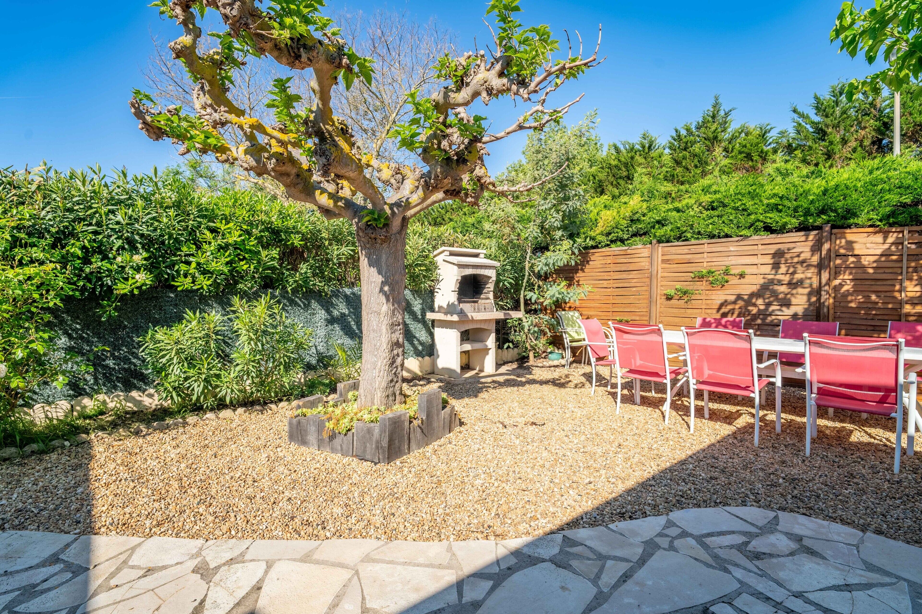 Eine Terrasse mit Steintisch, roten Stühlen, einem Baum und einem Grill.