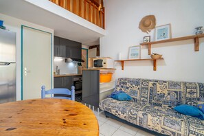 A compact living space with a dining table, a patterned sofa, a kitchen area, and a loft with wooden flooring.