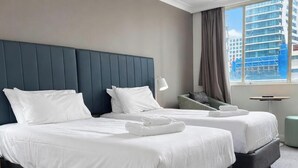 Une chambre d’hôtel avec deux lits, une tête de lit, une table de chevet, une lampe, une fenêtre donnant sur un immeuble et un petit pouf rond.