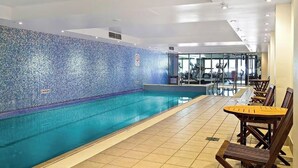 Une piscine aux carreaux de mosaïque bleus, un mur en verre et des chaises en bois disposées le long du bord.