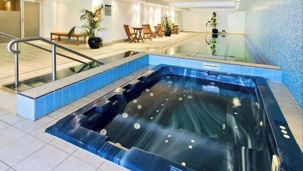 Une piscine intérieure moderne, dotée d’une paroi en verre, d’un jacuzzi et d’un espace détente avec des chaises et des tables.