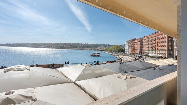 A terrace with umbrellas overlooking a waterfront with buildings in the background. - Room 'Luxury  Fronte Mare Con Jacuzzi' with Balcony, Wi-Fi and Air Conditioning (Napoli)