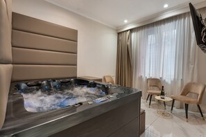 Una habitación moderna con una bañera de hidromasaje, dos sillones, una mesita con una botella y un vaso, y un televisor montado en la pared.