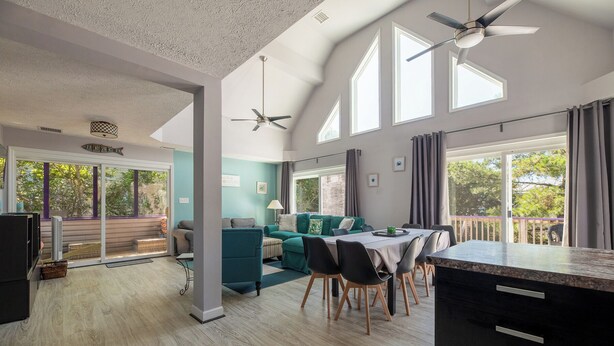 A spacious living area with a dining table, chairs, and a sofa. There are ceiling fans, large windows, and a view of the outdoors. - Lucky Fin - Corolla, Ocean Sands, Oceanside, 600 Yds to Beach, 4 Bedrooms, Hot Tub, Pets OK! (Corolla)