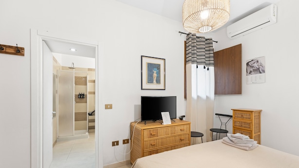 A bedroom with a bed, a television, a dresser, a wicker chair, a small table, and a doorway leading to a bathroom. - Room 'Camera 2' with Wi-Fi and Air Conditioning (Oristano)