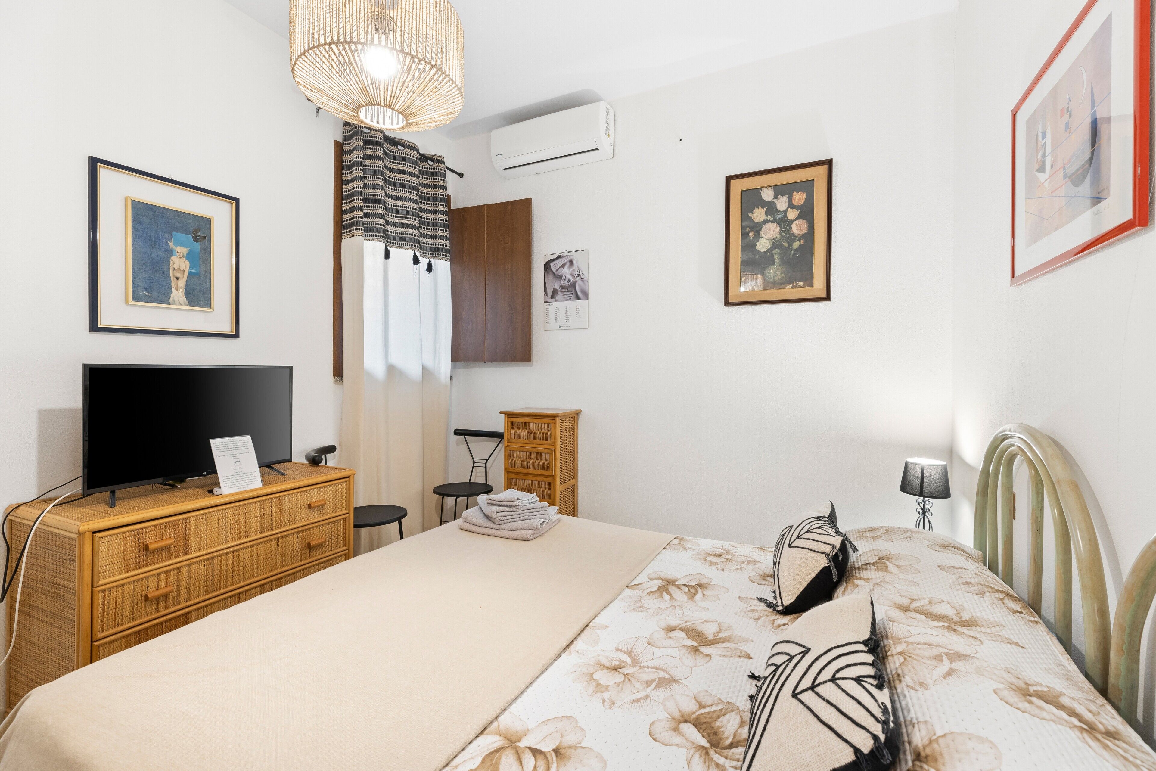A hotel room with a bed, a television, a wicker dresser, a wall-mounted air conditioner, and framed artwork.
