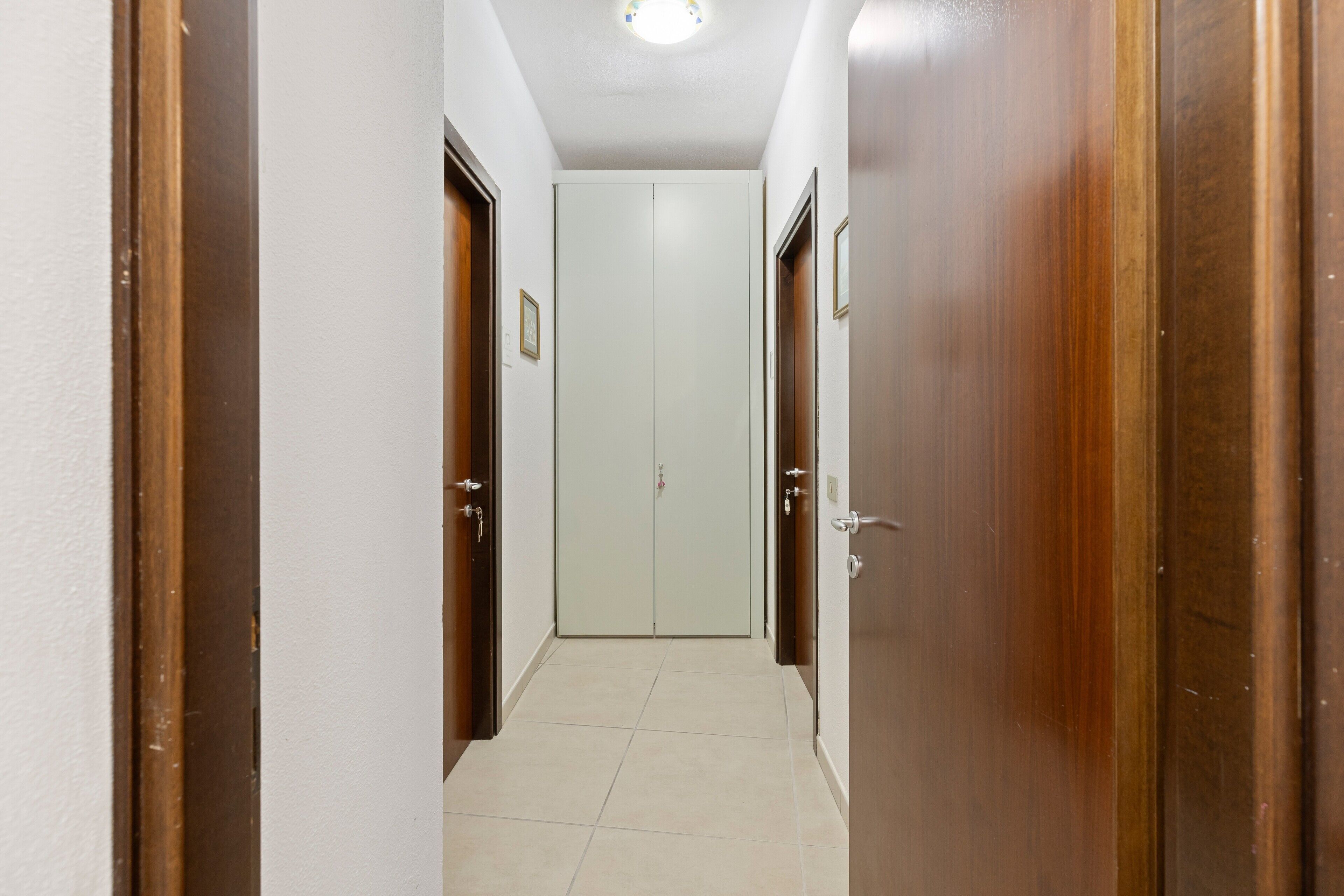 Un pasillo con una puerta al final, una lámpara en el techo y piso de baldosas.