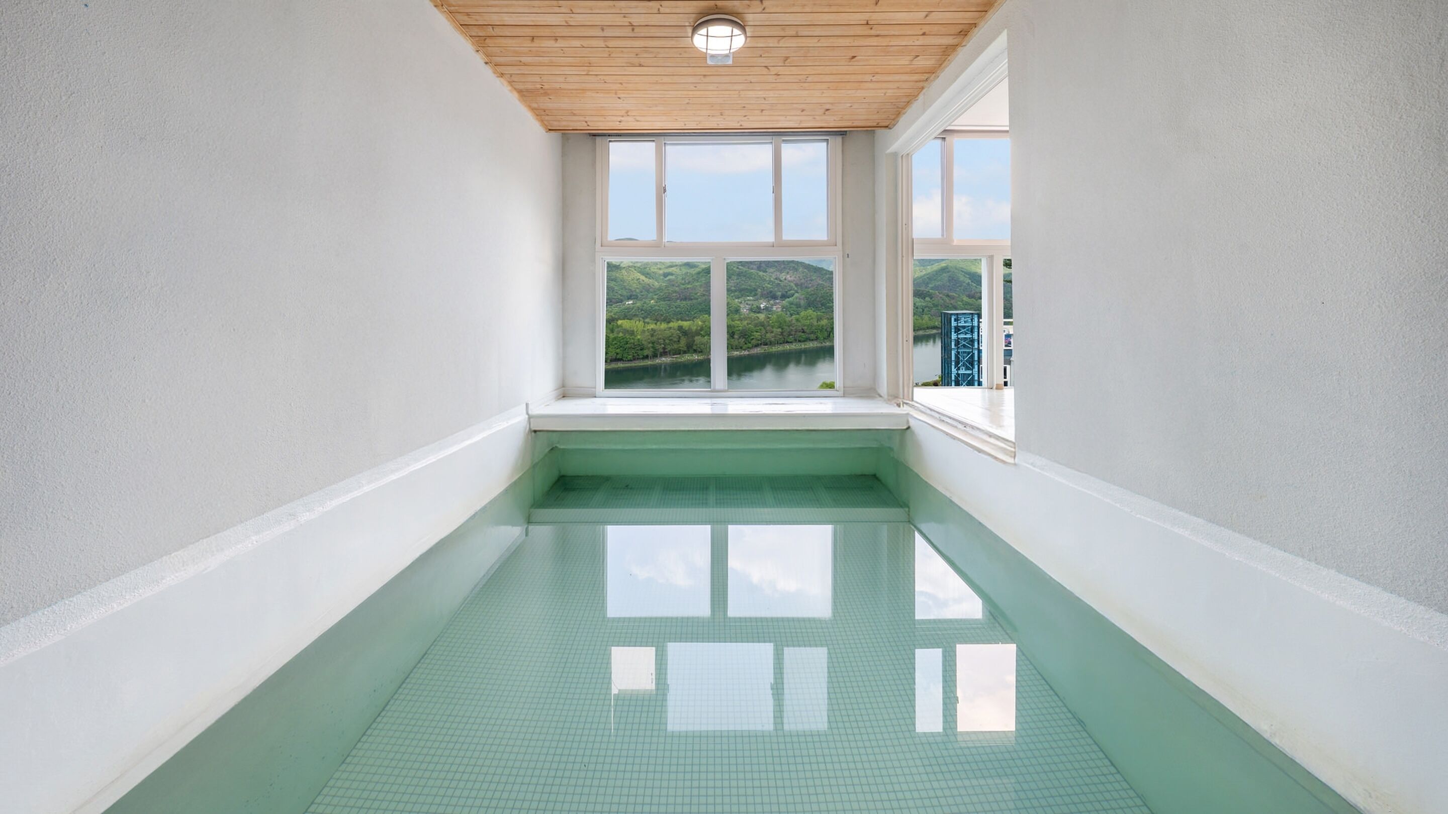 Ein Schwimmbecken mit Blick auf einen See und Hügel, umgeben von weißen Wänden und einer Holzbalkendecke.