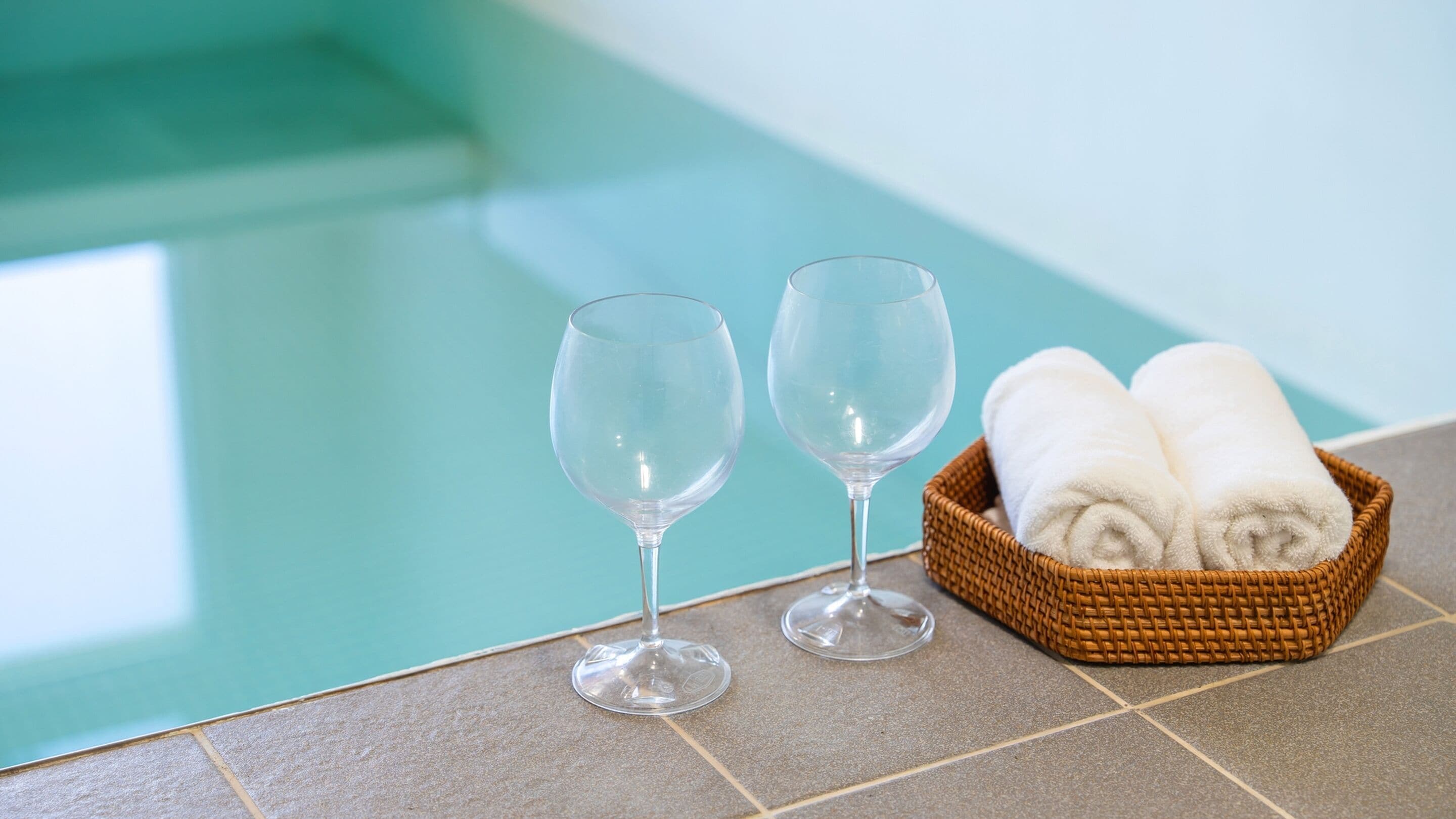 Due bicchieri da vino vuoti e un cesto di vimini con asciugamani arrotolati vicino a una piscina.