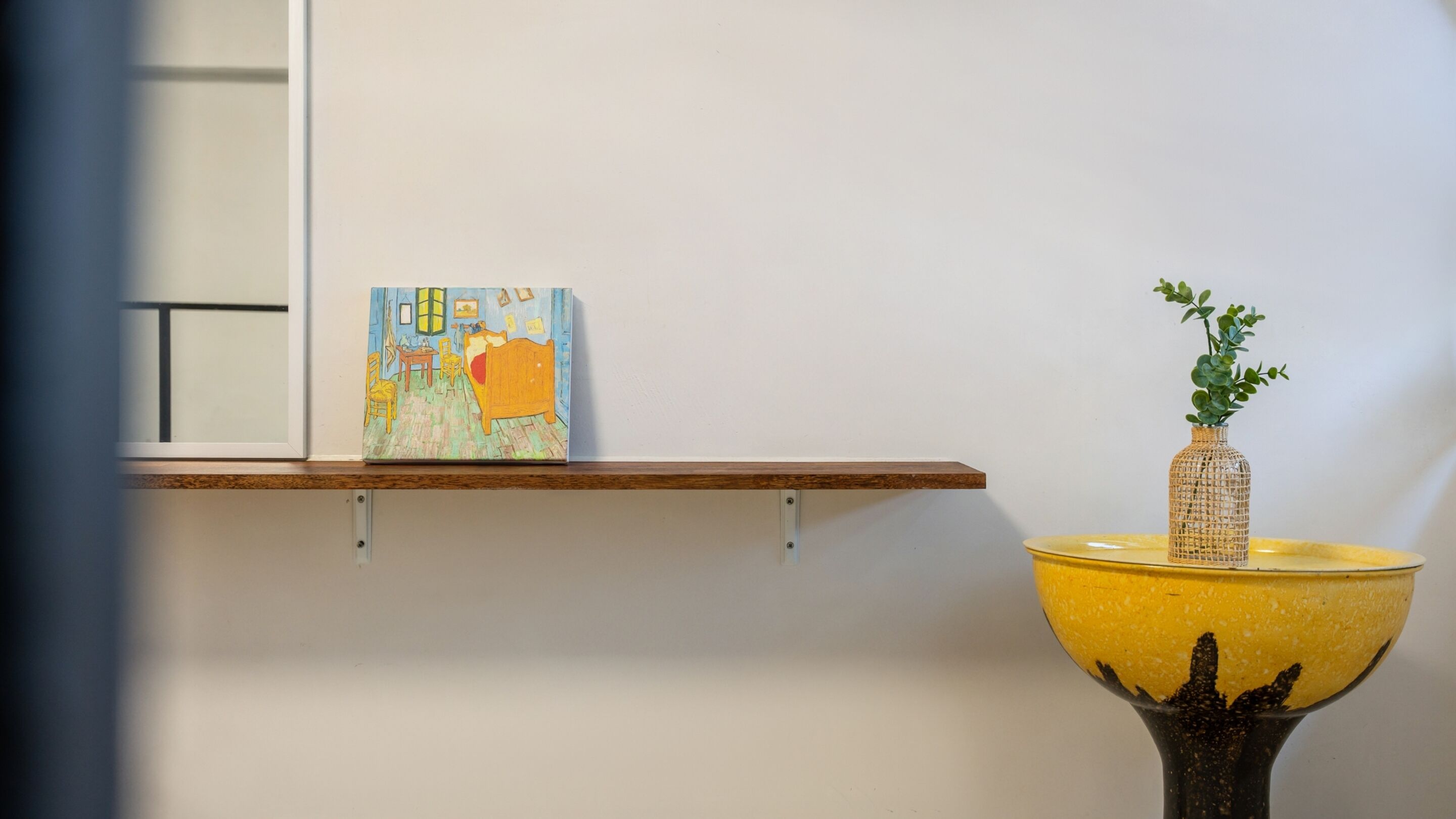 A room with a bed, a small table, and a shelf with a painting and a vase with a plant.