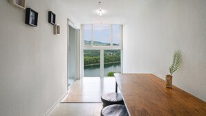 Une pièce au décor minimaliste, avec une table en bois et deux tabourets, une grande fenêtre donnant sur un lac et des collines, et une petite plante posée sur la table.