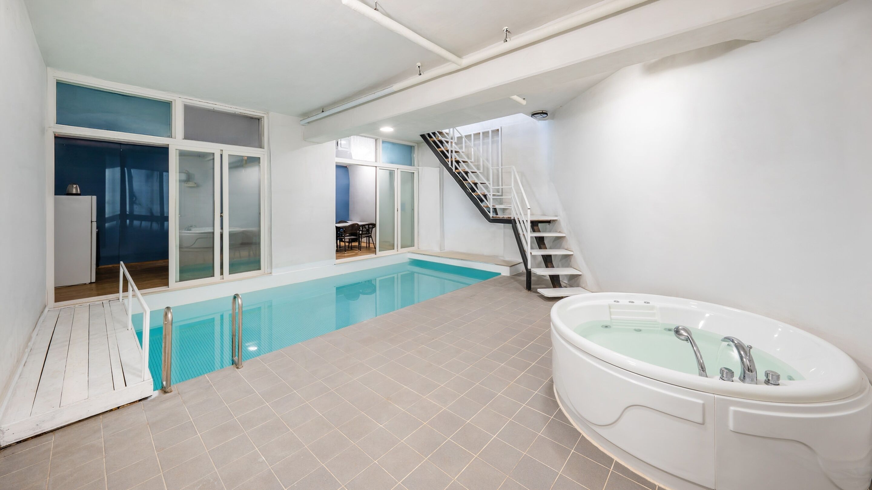 A modern indoor swimming pool with a glass-enclosed area, a separate jacuzzi, and a staircase leading to an upper level.