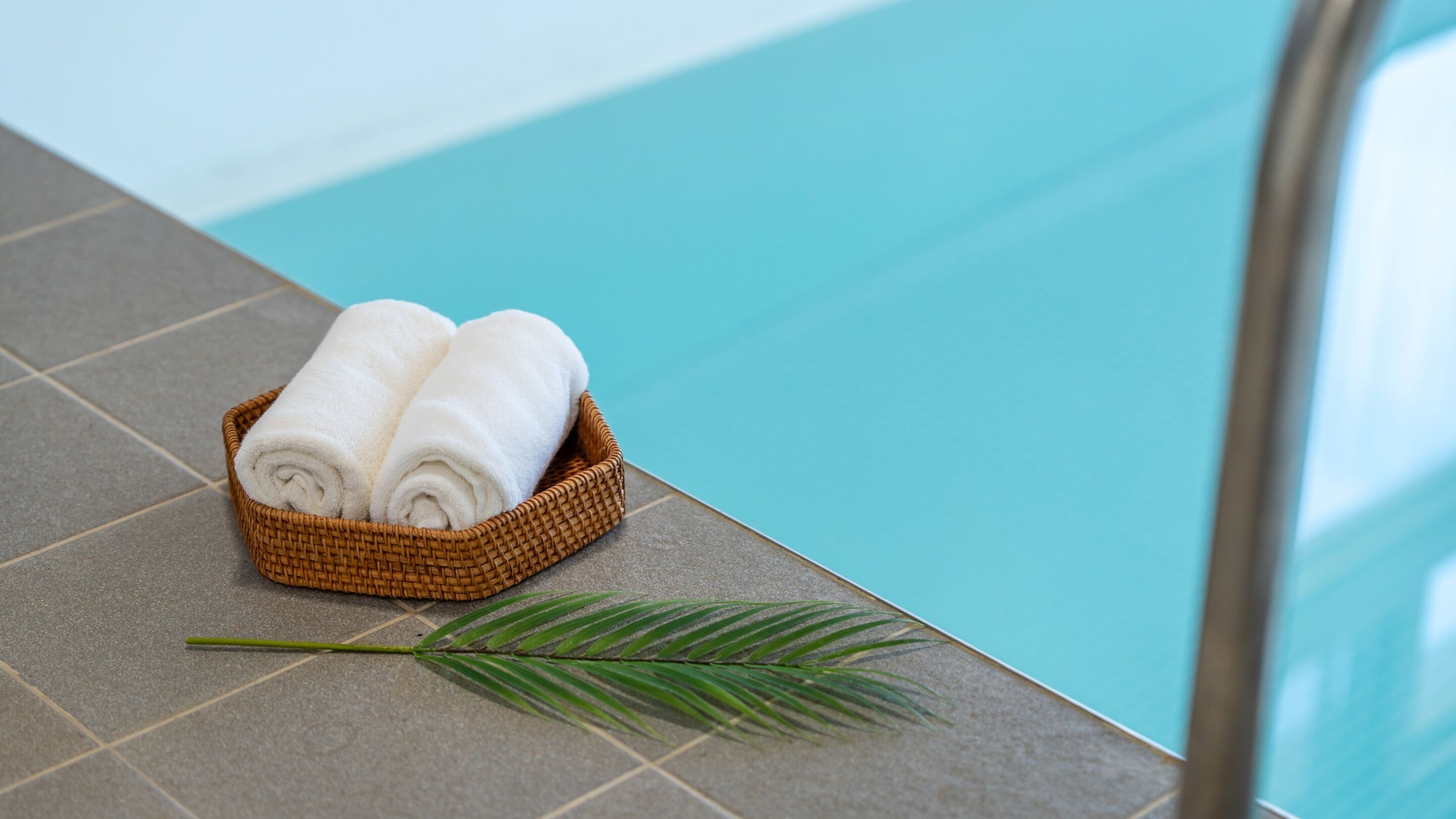 Un panier en osier contenant des serviettes blanches pliées, posé sur un sol carrelé, à côté d’une piscine.