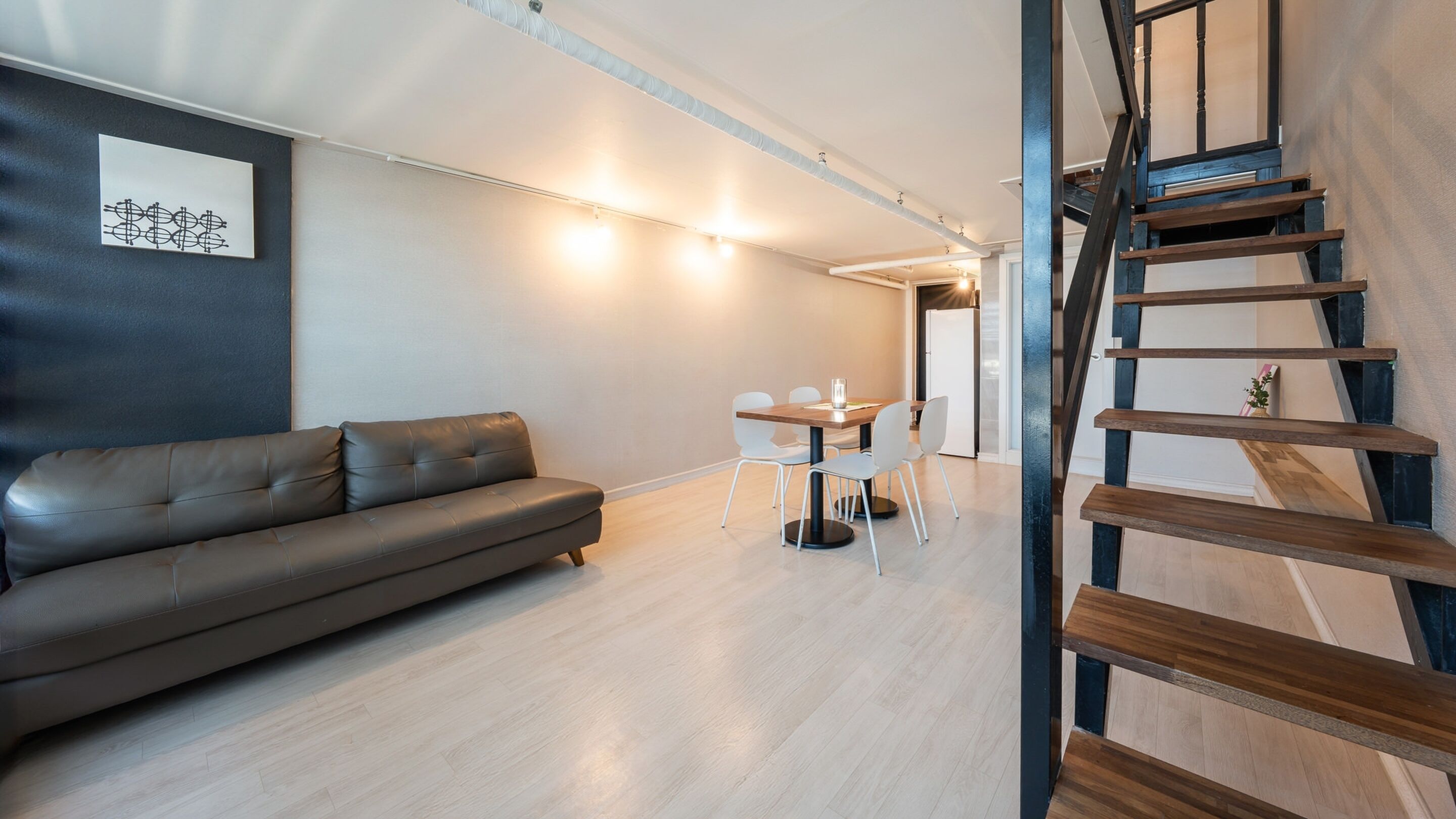A modern living room with a leather sofa, a dining table with chairs, and a wooden staircase.