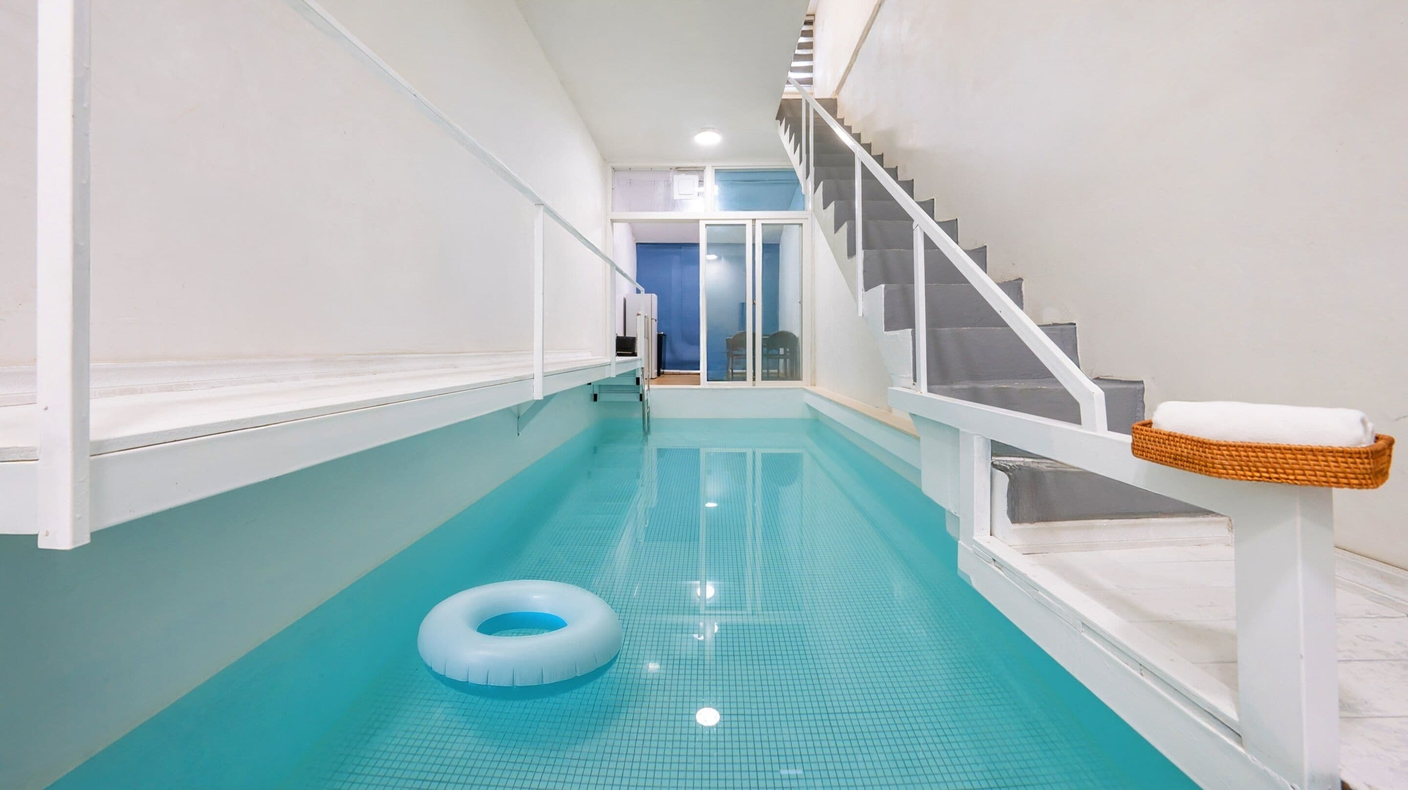 Une piscine intérieure étroite, dont le sol est recouvert de carreaux bleus, avec un flotteur blanc et un escalier orné d'un panier en osier sur la rambarde.