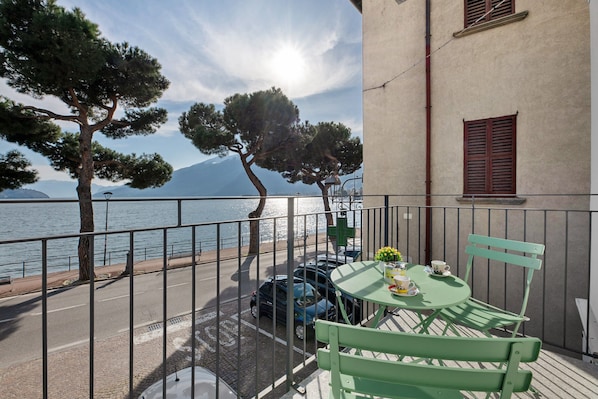 Un balcon avec une table et des chaises, donnant sur une rue et un plan d’eau, avec des arbres et des montagnes en arrière-plan.