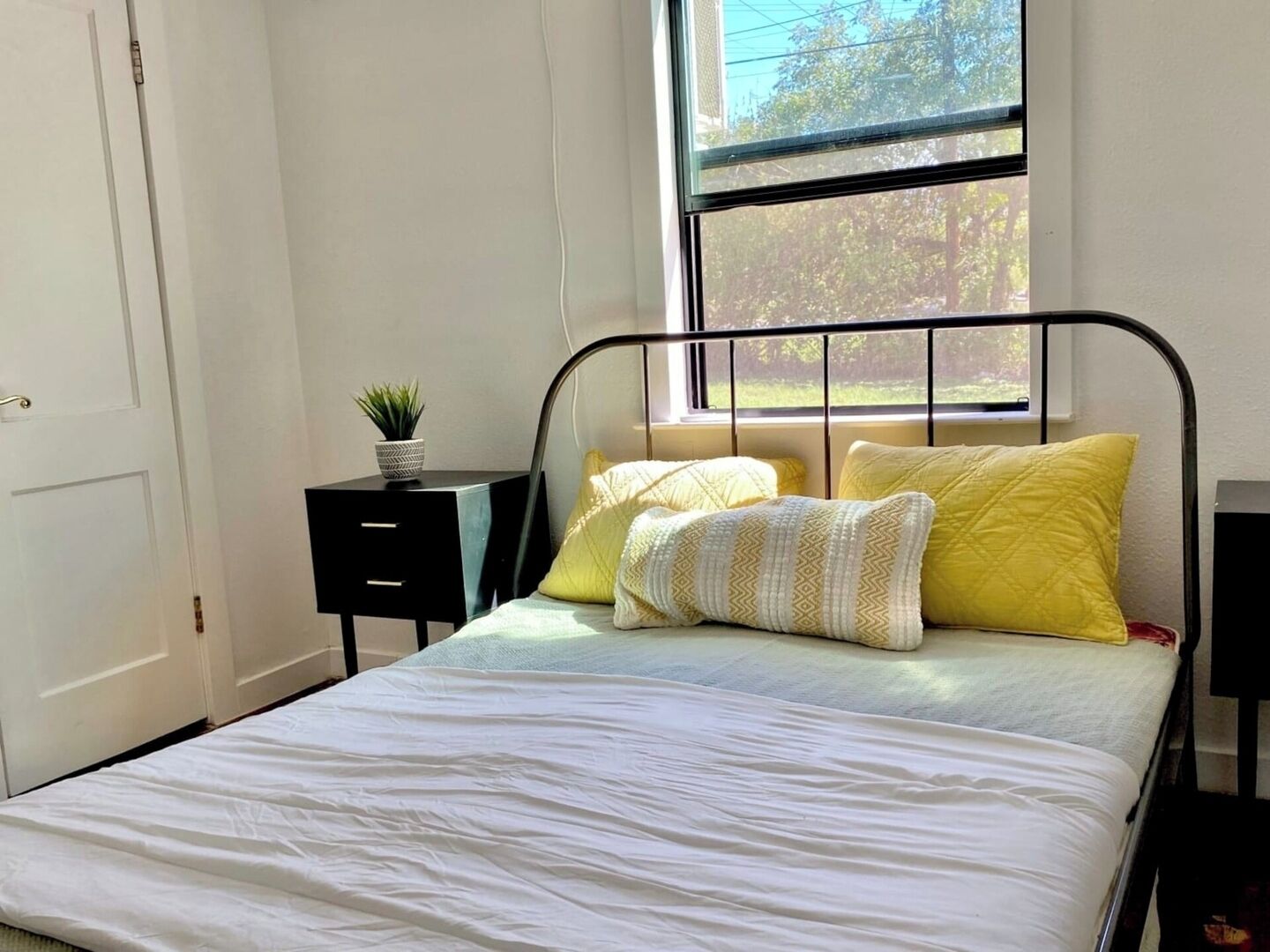 A bedroom with a bed, a window, a bedside table with a plant, and a small cabinet.