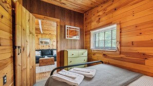 A wooden cabin room with a bed, a green dresser, a window with blinds, and a door leading to another room. - RC 17 – Cozy Cabin with Shared Rangeley Lake Waterfront, Kayaks, Paddleboards & Firepit (Rangeley)
