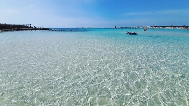 Une plage avec une eau turquoise et limpide, une côte lointaine et quelques personnes aperçues au loin.